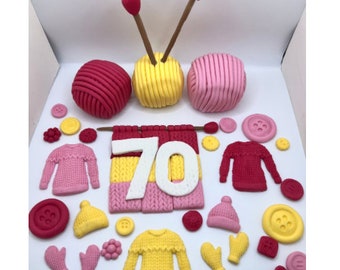 Könnte beinhalten: Ein rosa und gelber gestrickter Kuchentopper mit der Zahl 70 in der Mitte. Der Topper enthält Wollknäuel, Stricknadeln und andere Strick-Dekorationen.