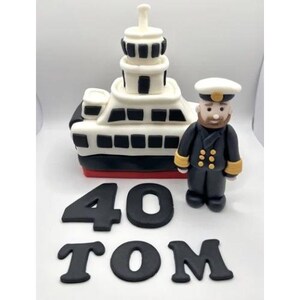 May include: Cake decoration with a white and black ship, red base, sailor figurine, and the name "TOM" and the number "40" in black. The ship has multiple levels and windows.