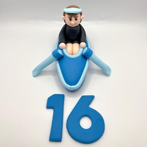 May include: A blue and white fondant cake topper of a person rowing a boat with the number 16 in blue fondant.