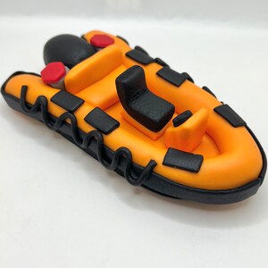 May include: A small, orange inflatable boat with black trim and a black seat. The boat has a rope-like detail around the edge.