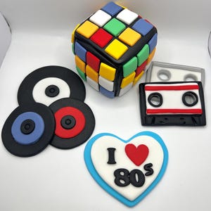 May include: A set of 80s themed cake toppers, including a Rubik's Cube, a cassette tape, two vinyl records, and a heart with the text "I love 80s".