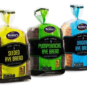 May include: Three loaves of Korn's Bakery Real Jewish Rye Bread. From left to right: Seeded Rye Bread, Pumpernickel Rye Bread, and Original Rye Bread. Each loaf is packaged in a clear plastic bag with a black and yellow, black and green, or black and blue label.