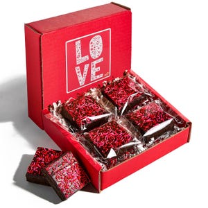 May include: A red box with the word "LOVE" in white, containing individually wrapped brownies with pink and red sprinkles. Two brownies are outside the box. The box is open, revealing the treats inside.