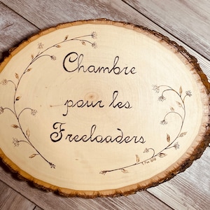May include: A wooden sign with a burnt-in design of a vine with flowers and the text "Chambre pour les Freeloaders".