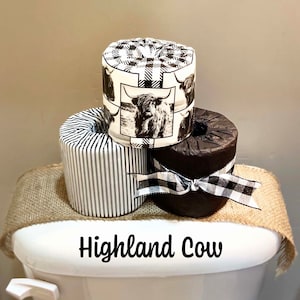 May include: Three rolls of toilet paper with different designs. The top roll has a black and white plaid pattern with a picture of a Highland cow. The middle roll has a black and white striped pattern. The bottom roll is brown with a black and white checkered ribbon. The rolls are sitting on a toilet tank with the words "Highland Cow" written on the front.