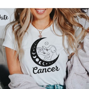 May include: White t-shirt with a black and white graphic of a woman with a crescent moon and stars. The text "Cancer" is printed below the graphic.