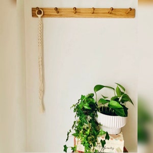 Peut inclure: Un porte-manteau mural en bois clair doté de six patères disposées horizontalement. Une longue corde en macramé blanc cassé, surmontée d'un anneau en bois, pend au premier crochet. Un cache-pot en céramique blanche côtelée contient des plantes.