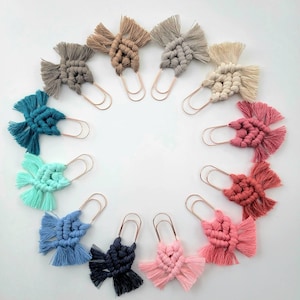 May include: A collection of twelve large paper clips, each featuring a decorative macrame knot design made from cotton cord in various colors including teal, blue, navy, pink, coral, beige, gray, and cream, arranged in a circular pattern.