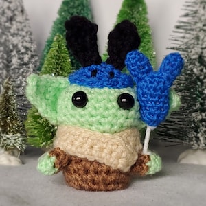 May include: A green crocheted character with a blue hat and reindeer antlers holding a blue balloon. The character is wearing a brown sweater and a beige scarf.