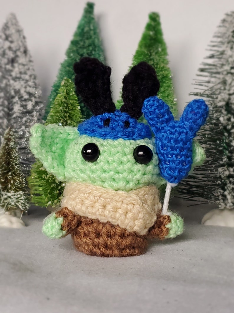 May include: A green crocheted character with a blue hat and reindeer antlers holding a blue balloon. The character is wearing a brown sweater and a beige scarf.
