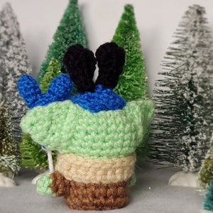 May include: A green crocheted character with blue ears and brown legs standing in front of a snowy background with miniature pine trees.