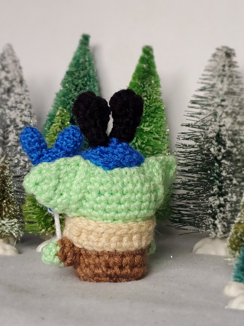 May include: A green crocheted character with blue ears and brown legs standing in front of a snowy background with miniature pine trees.