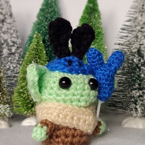 May include: A green crocheted amigurumi toy with black ears, a blue hat, and a blue crocheted hand holding a balloon. The toy is standing on a white surface with green trees in the background.