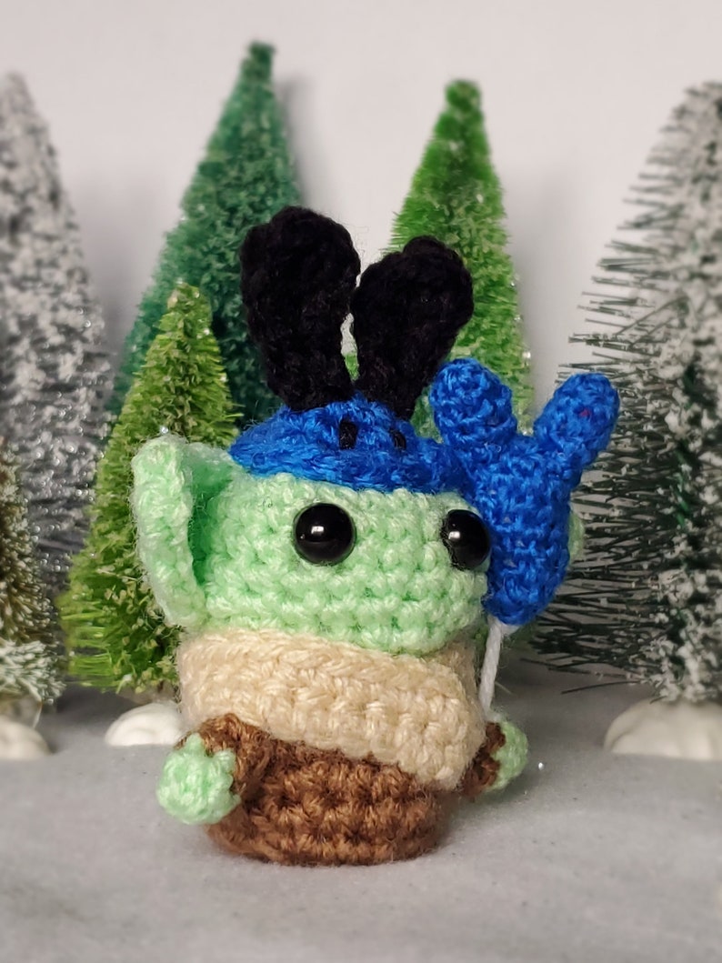May include: A green crocheted amigurumi toy with black ears, a blue hat, and a blue crocheted hand holding a balloon. The toy is standing on a white surface with green trees in the background.