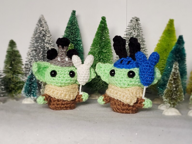 May include: Two green crocheted plush toys with large eyes and brown yarn clothing. One toy is holding a white crocheted balloon and the other is holding a blue crocheted balloon.