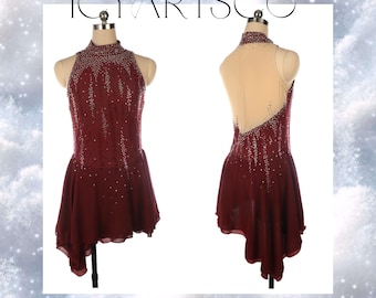 Peut inclure: Robe de patinage artistique bordeaux avec un col montant et un dos ouvert. Le corsage est orné de strass argentés. La jupe a un ourlet asymétrique. Le texte "ICYARTSCO" est en haut de l'image.