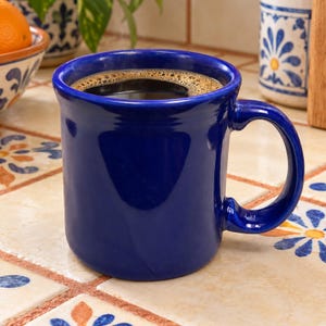 May include: A glossy, deep cobalt blue ceramic mug filled with dark black coffee. The mug features a thick, rounded rim and a sturdy, curved handle, showcasing a smooth, solid-colored finish that reflects light across its cylindrical body.