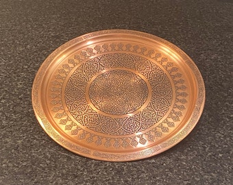 Puede incluir: Una bandeja redonda de cobre con un diseño intrincado en relieve. La bandeja tiene un borde elevado y un patrón circular en el centro.