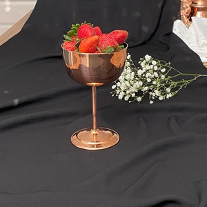 May include: A copper-colored metal vase with a tall stem and a round base, filled with fresh red strawberries. The vase is sitting on a black fabric surface.