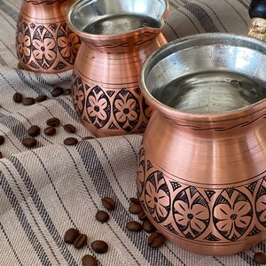 May include: Three copper coffee pots with ornate floral designs on a striped fabric background. The pots have wooden handles and are filled with water.