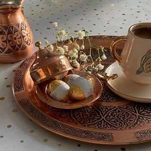 May include: A copper serving tray with intricate designs, holding a small copper bowl with three gold-wrapped chocolates. A white cup with a floral design and a saucer are on the right side of the tray. The tray is on a white tablecloth with gold polka dots.
