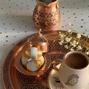 May include: A copper serving tray with intricate floral designs, featuring a small copper pot with a lid, a cup of coffee, and a plate with chocolate sweets. The tray is set on a white tablecloth with gold polka dots.
