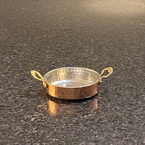 Könnte beinhalten: Eine kleine, runde Kupferpfanne mit gehämmerter Oberfläche und goldfarbenen Griffen. Die Pfanne ist leer und steht auf einer dunkelgrauen Oberfläche.