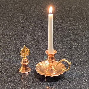 May include: A copper candle holder with a white candle lit inside. The candle holder has a decorative handle and a scalloped base. There is a small copper bell with a gold finial to the left of the candle holder.
