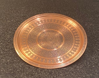 Puede incluir: Una bandeja redonda de servir de cobre con un diseño intrincado en relieve. La bandeja tiene un patrón circular con un círculo más pequeño en el centro.