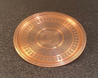 Puede incluir: Una bandeja redonda de servir de cobre con un diseño intrincado en relieve. La bandeja tiene un acabado de cobre oscuro y un borde elevado.