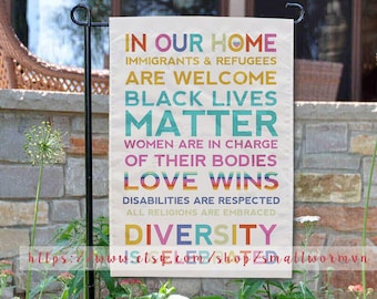 May include: A garden flag with a white background and colourful text that reads "In Our Home Immigrants & Refugees Are Welcome Black Lives Matter Women Are In Charge Of Their Bodies Love Wins Disabilities Are Respected All Religions Are Embraced Diversity".