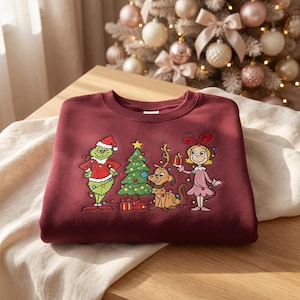 May include: A maroon crewneck sweatshirt featuring an embroidered graphic of a green character in a Santa suit, a decorated Christmas tree, a brown dog with antlers, and a blonde character in a pink dress, all wrapped in colorful string lights.