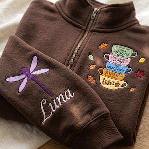 May include: A dark brown quarter-zip sweatshirt featuring an embroidered purple dragonfly and the name Luna on the sleeve. The chest displays a stack of five colorful mugs labeled Kim's Antiques, Stars Hollow Books, Dragonfly Inn, Al's Pancake World, and Luke's.