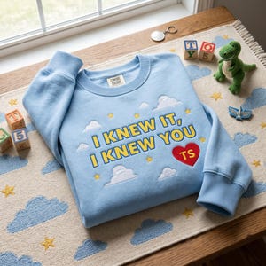 May include: A light blue crewneck sweatshirt featuring embroidered yellow text that reads I KNEW IT, I KNEW YOU, accompanied by a red heart with the letters TS, white clouds, and yellow stars. The garment has ribbed cuffs and a Comfort Colors label.