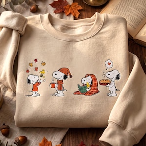 May include: A beige crewneck sweatshirt featuring a graphic print of Snoopy and Woodstock in four autumn-themed scenes: playing with falling leaves, holding a warm mug, reading under a plaid blanket, and carrying a freshly baked pie.