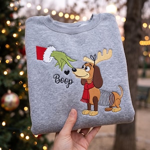 May include: A heather gray crewneck sweatshirt featuring an embroidered design of a green hand with a white cuff touching the nose of a brown dachshund dog wearing a red scarf and reindeer antlers. The word Boop is embroidered in black script.