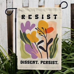 May include: A cream-colored garden flag with a colorful abstract floral design. The text "RESIST" is printed in black at the top of the flag. The text "DISSENT. PERSIST." is printed in black at the bottom of the flag.