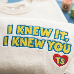 May include: A cream-colored sweatshirt featuring embroidered text that reads I KNEW IT, I KNEW YOU in yellow letters with blue outlines. Below the text is a red embroidered heart containing the yellow letters TS with blue outlines.
