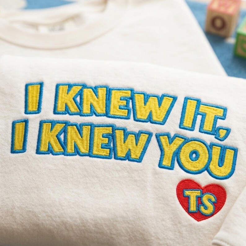 May include: A cream-colored sweatshirt featuring embroidered text that reads I KNEW IT, I KNEW YOU in yellow letters with blue outlines. Below the text is a red embroidered heart containing the yellow letters TS with blue outlines.