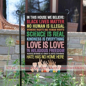 May include: A black garden flag with colorful text that reads "In this house we believe: Black Lives Matter, No Human is Illegal, Women's Rights are Human Rights, Science is Real, Kindness is Everything, Love is Love, In Religious Freedom, Injustice anywhere is a threat to justice everywhere, Hate has no home here". The flag also features a graphic of hands of different skin tones reaching up.