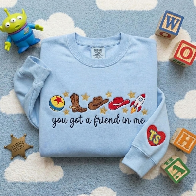 May include: A light blue crewneck sweatshirt featuring embroidered icons including a ball, cowboy boot, brown hat, red hat, and rocket ship, with the text "you got a friend in me" and a red heart on the sleeve containing the letters "TS".