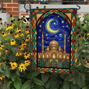 May include: A decorative garden flag featuring a stained-glass design of a mosque under a crescent moon and starry night sky. The flag has a colorful border with geometric patterns and is displayed on a black metal stand, surrounded by yellow flowers and green foliage.