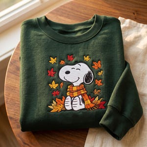 May include: A dark forest green crewneck sweatshirt featuring an embroidered graphic of a white beagle wearing a yellow and orange plaid scarf, surrounded by a variety of colorful autumn leaves in shades of red, orange, and yellow.