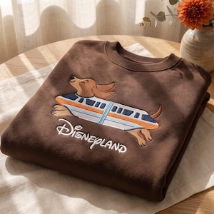 May include: A brown crewneck sweatshirt featuring an embroidered graphic of a dachshund dog wearing a monorail train costume. The design includes the word Disneyland in white script lettering below the dog. The sweatshirt has a ribbed collar.