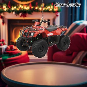 May include: A red ATV ornament with black tires and a black seat is suspended from a chain. The ATV is decorated with colorful Christmas lights. The text "IMANDA" is visible on the side of the ATV. The words "Clear Acrylic" are in the upper right corner.