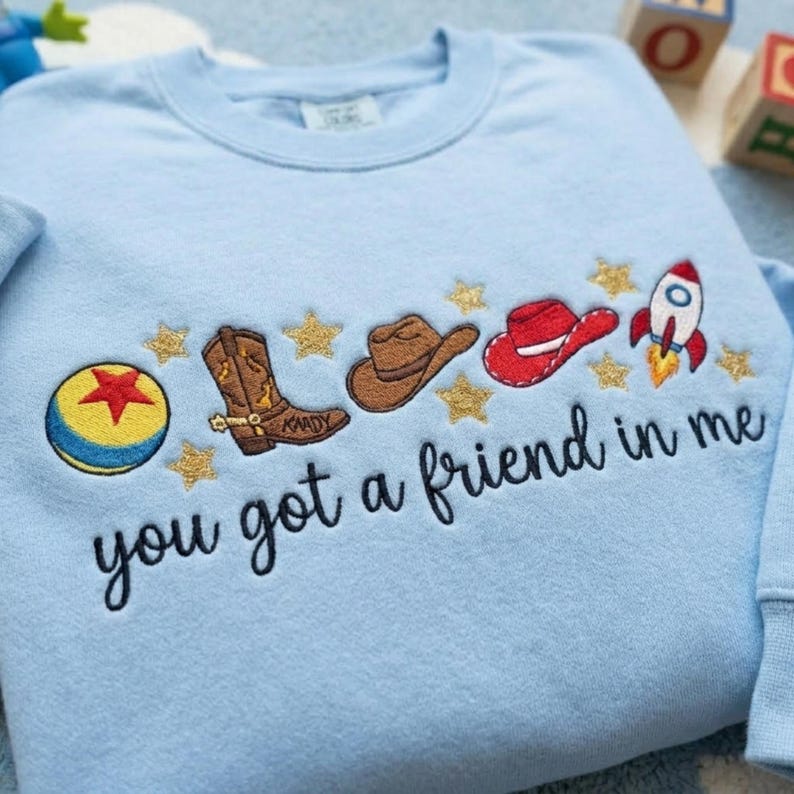 May include: A light blue crewneck sweatshirt featuring embroidered designs of a yellow and blue ball with a red star, a brown cowboy boot, a brown cowboy hat, a red hat, and a rocket ship, interspersed with gold stars and the text you got a friend in me.