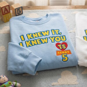 Puede incluir: Una sudadera de cuello redondo azul claro con texto bordado en amarillo que dice I KNEW IT, I KNEW YOU. Debajo del texto hay un coraz&oacute;n rojo con las letras TS, un rect&aacute;ngulo naranja con la palabra STORY y un n&uacute;mero 5 grande en amarillo.