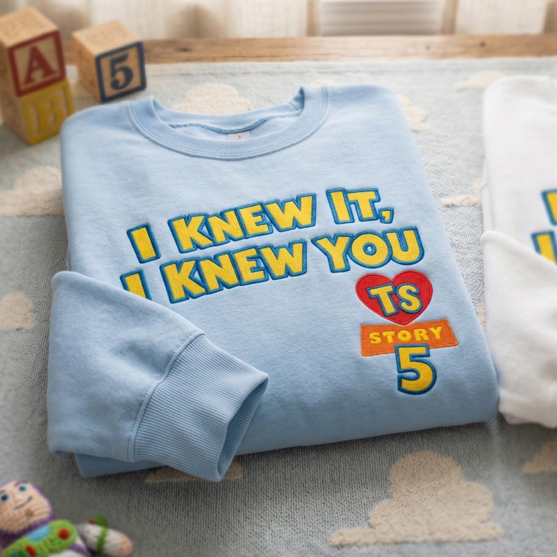 Puede incluir: Una sudadera de cuello redondo azul claro con texto bordado en amarillo que dice I KNEW IT, I KNEW YOU. Debajo del texto hay un coraz&oacute;n rojo con las letras TS, un rect&aacute;ngulo naranja con la palabra STORY y un n&uacute;mero 5 grande en amarillo.