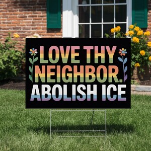 May include: A black yard sign with the words "LOVE THY NEIGHBOR. ABOLISH ICE" in rainbow-colored letters. The sign is supported by a white metal frame and is placed on a green lawn in front of a brick house.