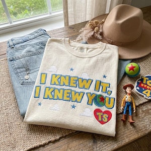 K&ouml;nnte beinhalten: Ein cremefarbenes T-Shirt mit gesticktem Text, der I KNEW IT, I KNEW YOU in Gelb mit blauen Umrandungen zeigt. Das Design enth&auml;lt kleine blaue Sterne, wei&szlig;e Wolken, einen bunten Ball mit einem roten Stern und ein rotes Herz mit den Buchstaben TS.